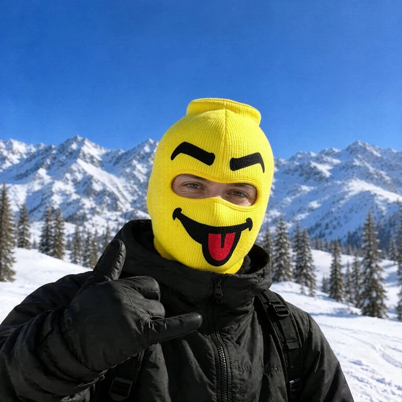 Blep yellow big head ski mask with a cheeky grin and red tongue out, worn on a snowy mountain
